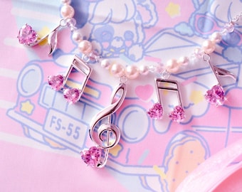May include: A delicate necklace featuring musical note charms and a treble clef pendant. The charms are adorned with pink heart-shaped gemstones and are strung on a strand of pearls. The background is a soft pink with a cartoon design.