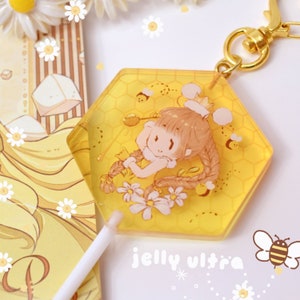 Op de afbeelding: Een gele zeshoekige sleutelhanger met een cartoonillustratie van een meisje met een kroon, omringd door bijen en bloemen. De sleutelhanger heeft een goudkleurige sluiting. De tekst "jelly ultra" is zichtbaar.