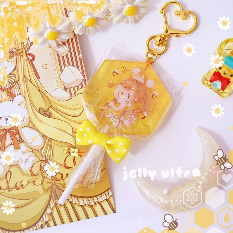 Op de afbeelding: Een verzameling geel-thema accessoires en briefpapier. Inclusief een zeshoekige sleutelhanger met een cartoonfiguur, een lolly met een gele strik, een halve maan decoratie en een wenskaart met een teddybeer illustratie. De tekst "jelly ultra" is zichtbaar.