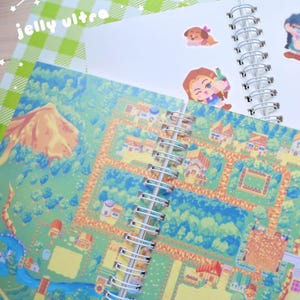 May include: Spiral-bound notebooks with colorful, illustrated pages. One notebook features a detailed map design with houses, trees, and a river. The other has white pages with stickers of cartoon characters. The text "jelly ultra" is visible.