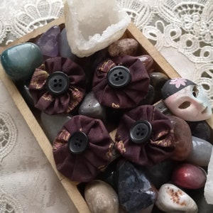 May include: Four brown fabric flower-shaped hair clips with black buttons in the center. The clips are arranged in a wooden box with various stones and crystals.