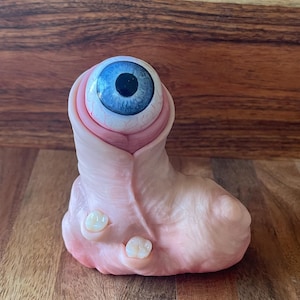 Könnte beinhalten: Eine skurrile Skulptur mit einem realistischen blauen Auge mit schwarzer Pupille, eingebettet in eine fleischfarbene, rosa-beige Form. Zwei kleine, weiße Zähne sind ebenfalls vorhanden. Das Design ist ungewöhnlich und auffällig.