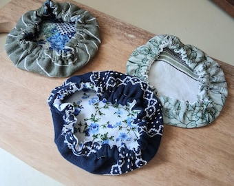 May include: Three circular fabric bowl covers with elasticized edges. One cover is olive green with white stripes, one is navy blue with a white floral and geometric pattern, and one is light green with a dark green botanical leaf print.