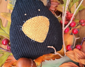 May include: A black ribbed fabric ornament with a triangular yellow patterned patch stitched onto the front. The ornament features two small light-colored dots for eyes and a dark string at the top, nestled among artificial autumn leaves and berries.
