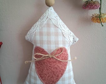 May include: A house-shaped fabric ornament featuring a beige and white gingham pattern, accented with white lace trim along the roofline. A red heart with a floral pattern is stitched to the center, tied with a jute twine bow and a wooden bead.