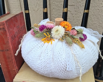 May include: A white fabric pumpkin covered in a delicate lace overlay, topped with a cluster of dried flowers in shades of yellow, orange, pink, and purple. A small wooden cylinder serves as the stem, and the pumpkin rests near two vintage books.