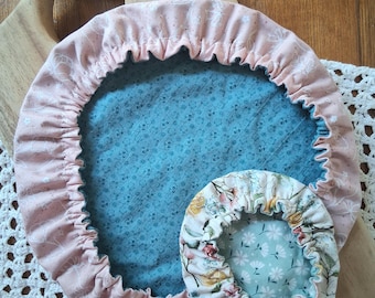 May include: Two round fabric bowl covers with elasticized edges. The larger cover features a dusty pink exterior with a white line-drawn floral pattern and a teal interior with a small white floral print. The smaller cover has a floral print.