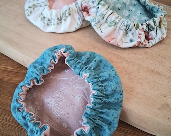 May include: Three round fabric bowl covers with elasticized edges. One cover features a white background with green leaf patterns and a pink interior. Another has a floral print with blue and pink tones. The third is teal with a pink floral interior.