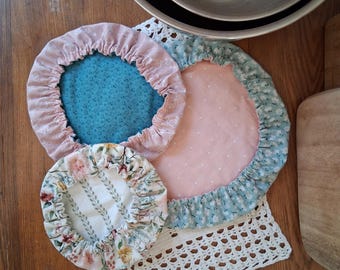 May include: Three round fabric bowl covers with elasticized edges in various patterns including pink, teal, and floral prints. The set includes a small floral cover, a medium teal cover with a pink border, and a large pink cover with a blue floral border.
