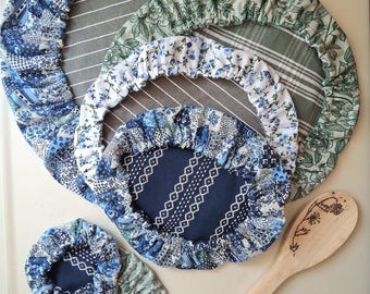 May include: A collection of five round fabric bowl covers in varying sizes, featuring blue and white patchwork, green floral, and striped patterns with elasticized edges, accompanied by a light-colored wooden spoon with a floral wood-burned design.