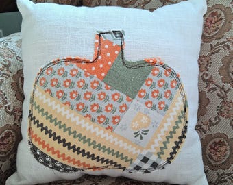 May include: A square white throw pillow featuring a central pumpkin applique made from various fabric patches. The pumpkin design includes orange floral, green, yellow chevron, and black and white gingham patterns with dark stitched outlines.