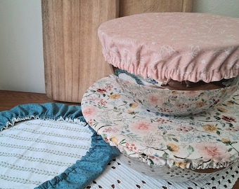 May include: Three circular fabric bowl covers with elasticized edges. One cover is solid dusty pink with a white line-drawn floral pattern. Another features a colorful floral print with roses. The third is teal with a white leaf pattern.