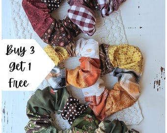 May include: Three fabric hair scrunchies featuring patchwork designs. The top scrunchie has maroon, brown, and white patterns including polka dots and gingham. The middle one features yellow honeycomb, orange, and white prints. The bottom one displays green, floral, and brown polka dot patterns.