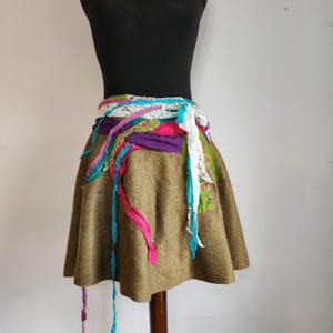 May include: A brown flared skirt with a multi-coloured sash. The sash includes purple, pink, green, and blue ribbons, and white lace. The skirt is displayed on a mannequin.