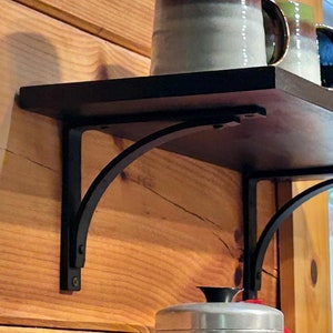 May include: A black metal shelf bracket with a curved design is mounted to a wooden wall. The shelf is made of black wood and has a variety of mugs and a silver container on top.