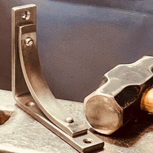 K&ouml;nnte beinhalten: Eine silberne Metall-Regalhalterung mit gebogenem Design, die auf einer Metalloberfl&auml;che neben einem schwarzen Hammer liegt.