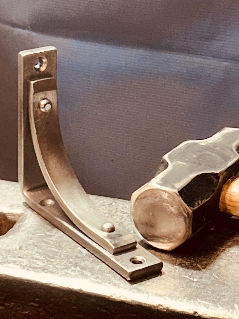 K&ouml;nnte beinhalten: Eine silberne Metall-Regalhalterung mit gebogenem Design, die auf einer Metalloberfl&auml;che neben einem schwarzen Hammer liegt.