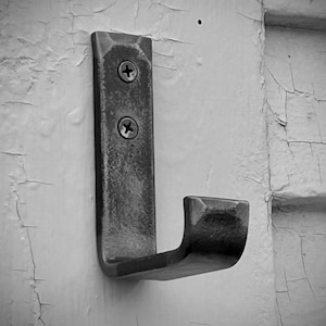 May include: A black metal hook with two screws attached to a white wall. The hook is bent at a 90-degree angle, forming a J-shape.