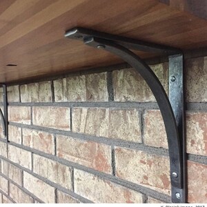 May include: A pair of black metal shelf brackets with a curved design, mounted to a brick wall and supporting a wooden shelf.