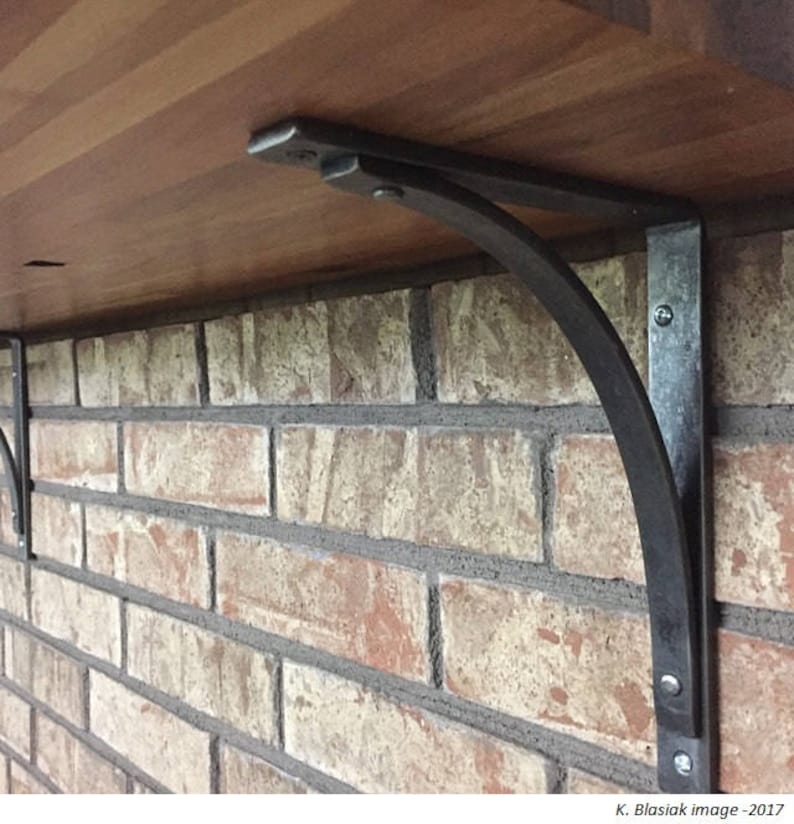 May include: A pair of black metal shelf brackets with a curved design, mounted to a brick wall and supporting a wooden shelf.