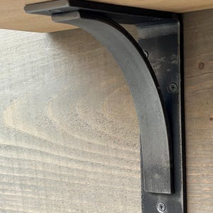 May include: Black metal shelf bracket with a curved design. The bracket is attached to a wooden surface.