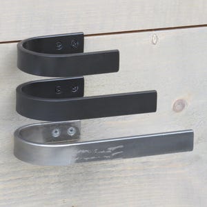 May include: Three metal towel bars, two black and one silver, are mounted on a white wooden wall. The bars are curved and have a simple design.