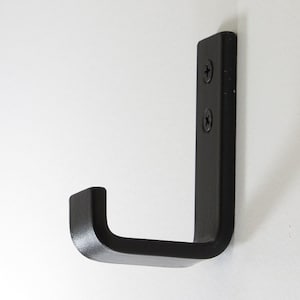 May include: Black metal hook with a rounded edge. The hook is mounted to a white wall.