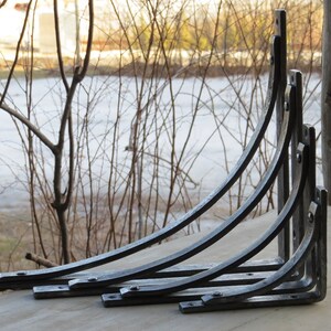 May include: Set of five black metal shelf brackets, arranged in size order, with the largest bracket at the top and the smallest at the bottom. The brackets are curved and have a decorative design.