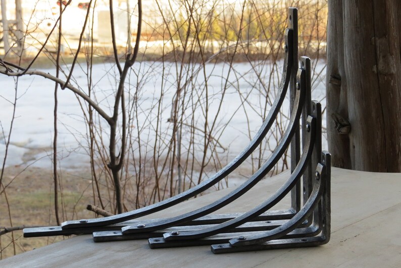 May include: Set of five black metal shelf brackets, arranged in size order, with the largest bracket at the top and the smallest at the bottom. The brackets are curved and have a decorative design.