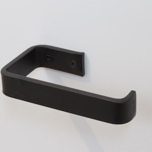 May include: A matte black metal toilet paper holder with a flat, rectangular profile. The holder features a U-shaped design with a mounting plate that attaches to the wall using two visible black screws, providing a simple and functional fixture.