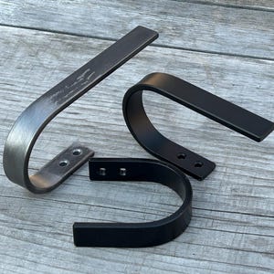 May include: Two metal shelf brackets, one in a brushed silver finish and one in black. Both brackets have a curved shape and are designed to be mounted to a wall.