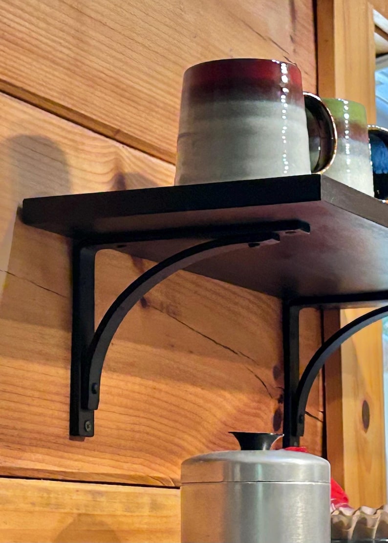May include: A black metal shelf bracket with a curved design is mounted to a wooden wall. The shelf is made of black wood and has a variety of mugs and a silver container on top.
