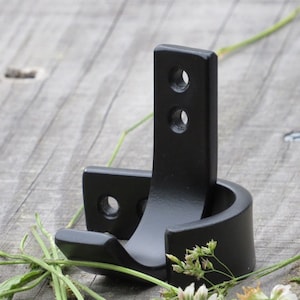 May include: Black metal hook with two mounting holes. The hook is designed to hold a round object, such as a broom or a mop.