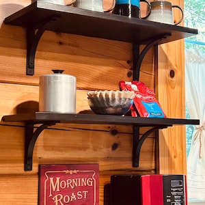 May include: Two black shelves with metal brackets, holding mugs, a silver container, a bowl, and a bag of coffee. The shelves are mounted on a wood wall. A red coffee maker is below the shelves.