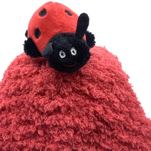 Puede incluir: Un juguete de peluche de mariquita roja con lunares negros y una cabeza negra. El juguete tiene un cuerpo suave y texturizado y está diseñado para ser tierno. La cara de la mariquita tiene ojos bordados y una boca sonriente.