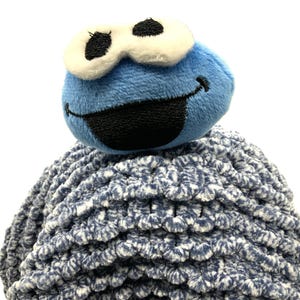 May include: Blue plush toy with a smiling face and large white eyes. The toy is sitting on top of a blue and white fuzzy ball.