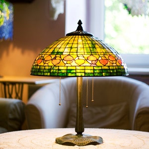 May include: A stained glass table lamp with a green, yellow, and orange design. The lamp is on a white table with a crocheted tablecloth.