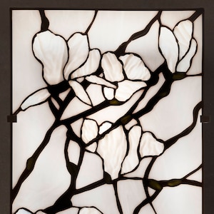 Puede incluir: Un panel de vitral con un diseño de flores blancas y ramas negras. El panel está enmarcado en metal negro y está de pie sobre una superficie oscura.