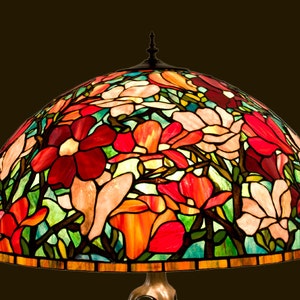May include: A stained glass lampshade with a floral design featuring pink, red, orange, and green glass pieces. The shade is dome-shaped and has a bronze base.