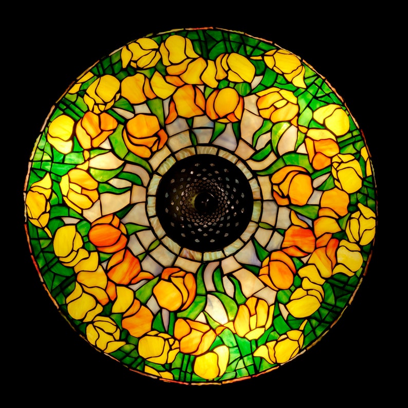 May include: A stained glass lampshade with a circular design featuring yellow, green, and orange tulips. The lampshade is illuminated from within, creating a warm glow.