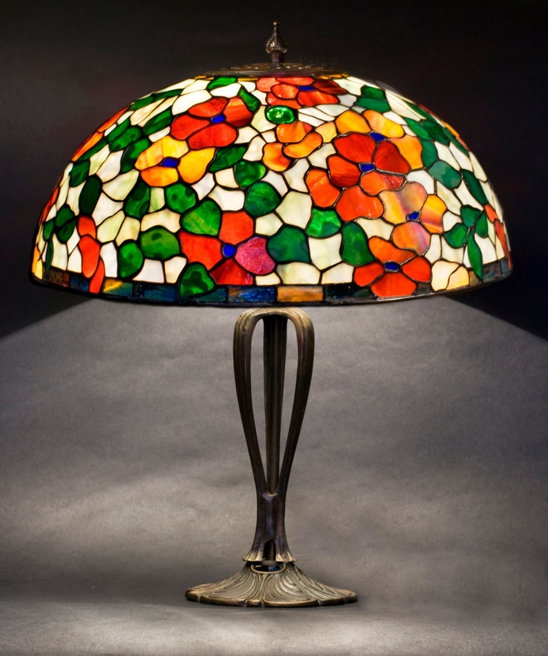 May include: A stained glass table lamp with a floral design in shades of red, orange, green, and white. The lamp has a bronze base with a decorative design.