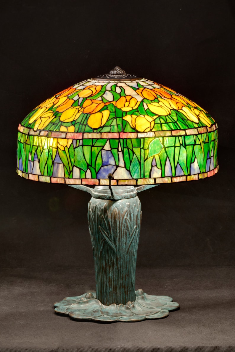 May include: A stained glass table lamp with a green and yellow tulip design. The base is a bronze green color and has a leaf design.