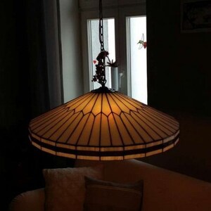 Puede incluir: Una l&aacute;mpara de techo de cristal tintado con un marco de metal marr&oacute;n y una pantalla de vidrio beige. La luz cuelga de una cadena y tiene un peque&ntilde;o p&aacute;jaro decorativo en la parte superior.