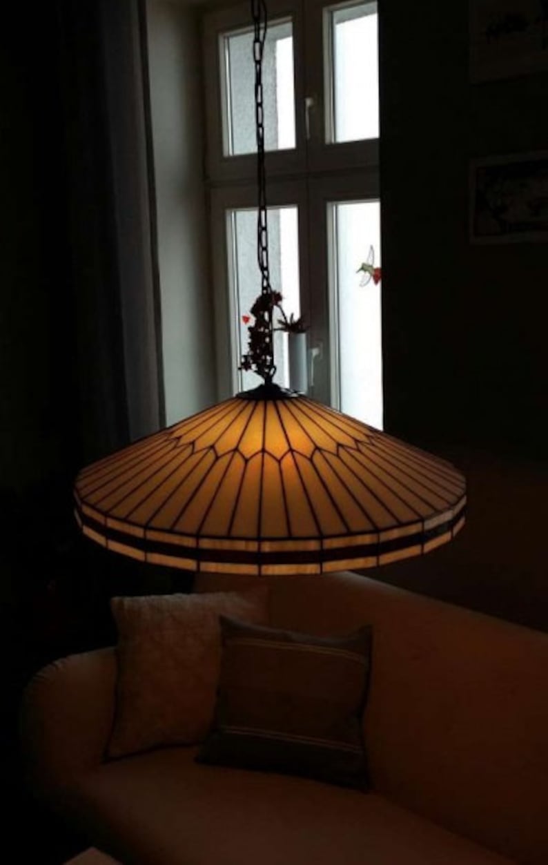 Puede incluir: Una l&aacute;mpara de techo de cristal tintado con un marco de metal marr&oacute;n y una pantalla de vidrio beige. La luz cuelga de una cadena y tiene un peque&ntilde;o p&aacute;jaro decorativo en la parte superior.