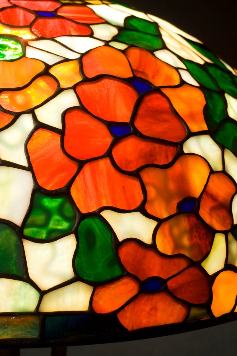 May include: A stained glass lampshade with a floral design. The shade features red, orange, green, and white glass pieces. The glass is cut into various shapes and sizes, creating a unique and intricate pattern.