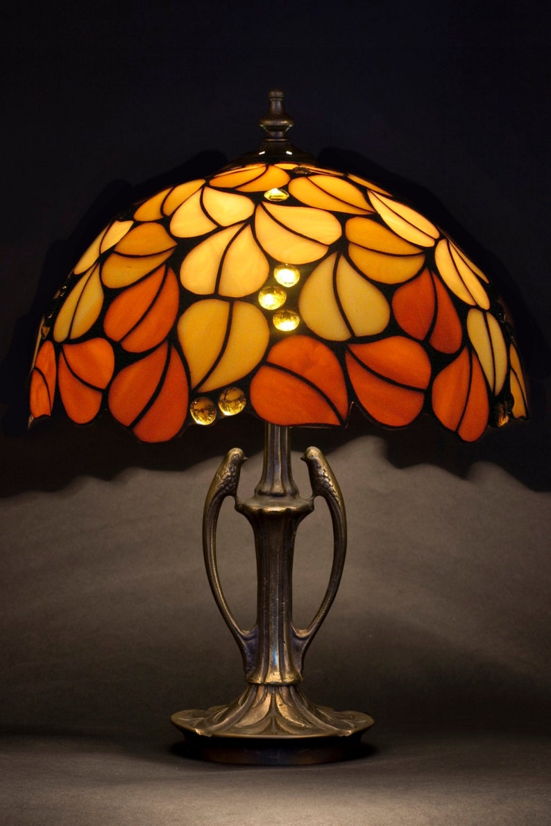 Puede incluir: Una l&aacute;mpara de mesa de vidrio emplomado con un dise&ntilde;o floral naranja y amarillo. La l&aacute;mpara tiene una base de bronce con un dise&ntilde;o decorativo.