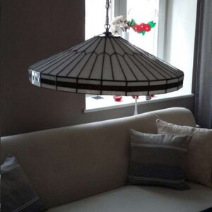 Puede incluir: Una l&aacute;mpara de techo de cristal blanco con un marco de metal negro cuelga de una cadena. La l&aacute;mpara est&aacute; en una habitaci&oacute;n con un sof&aacute; blanco y almohadas.
