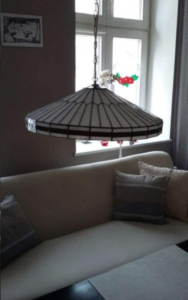 Puede incluir: Una l&aacute;mpara de techo de cristal blanco con un marco de metal negro cuelga de una cadena. La l&aacute;mpara est&aacute; en una habitaci&oacute;n con un sof&aacute; blanco y almohadas.