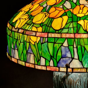May include: A stained glass lampshade with a floral design featuring yellow, orange, and green tulips. The lampshade is mounted on a bronze base with a textured surface.