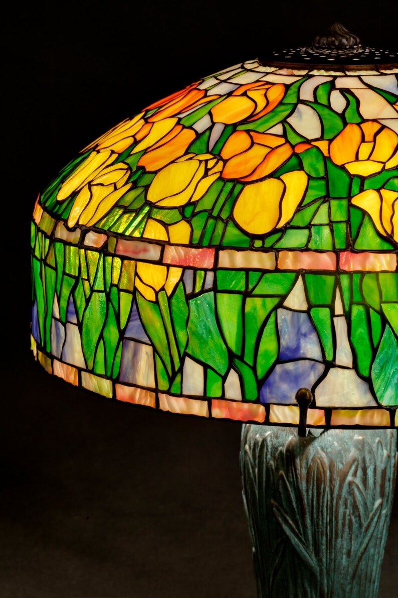 May include: A stained glass lampshade with a floral design featuring yellow, orange, and green tulips. The lampshade is mounted on a bronze base with a textured surface.